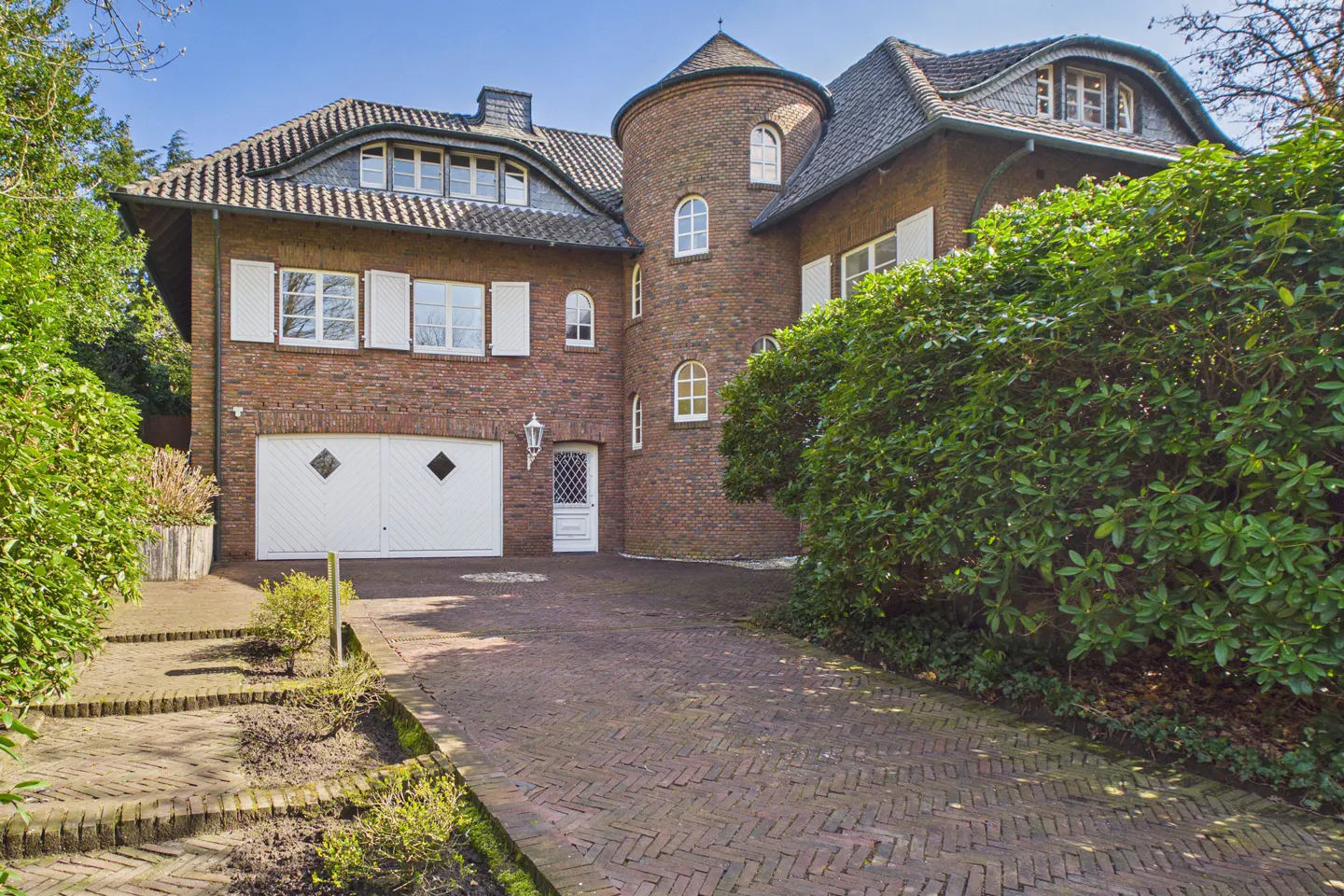 Brick house with a turret, white trim, and a brick driveway. Green bushes surround the house.