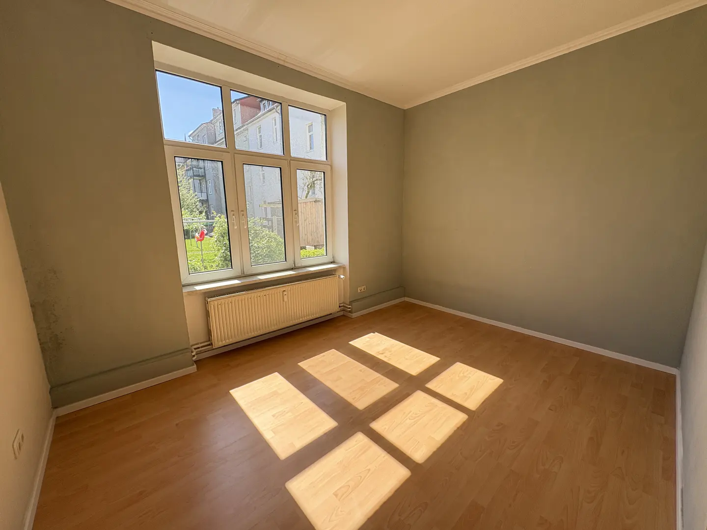 Empty room with light green walls, wood floor, and a large window with a radiator underneath. Sunlight streams in, creating bright squares on the floor.