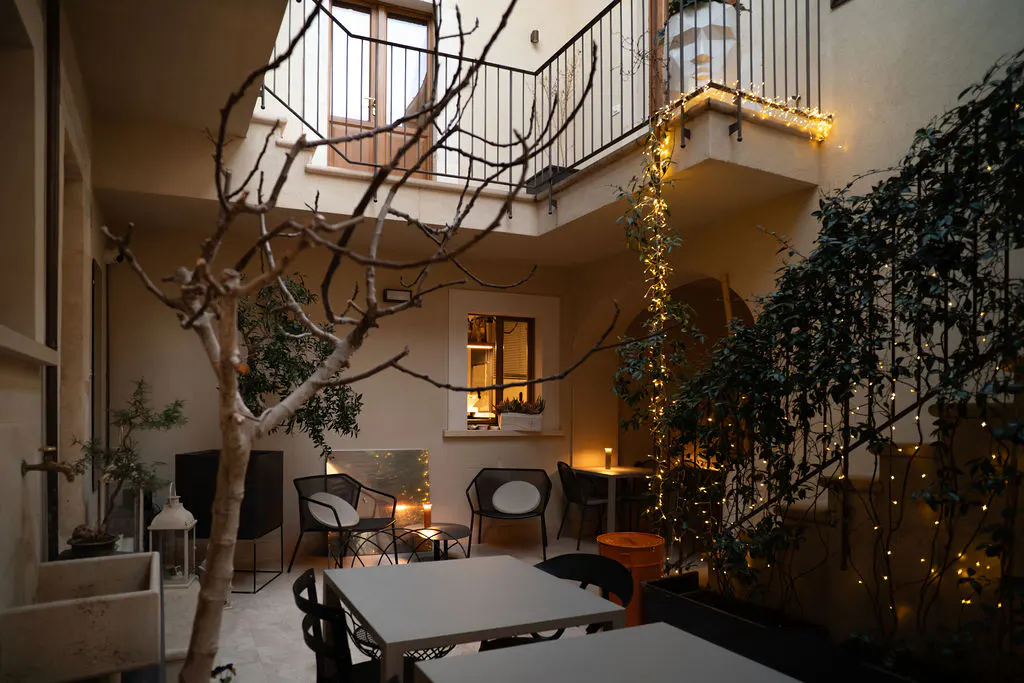 Cozy courtyard with tables, chairs, and plants. Balcony with lights above. Tree in foreground. Warm, inviting atmosphere.