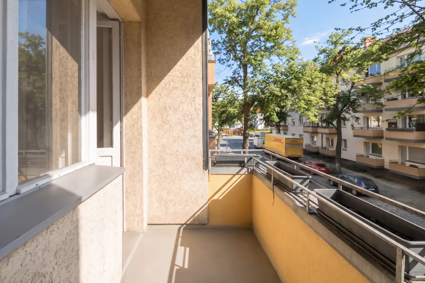 Balcony view with white window frame, beige walls, and metal railing overlooking a street with cars and a yellow DHL truck.