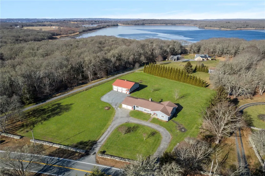 Aerial view of a light blue house with a red-roofed garage on a large green lawn near a body of water and a forest.