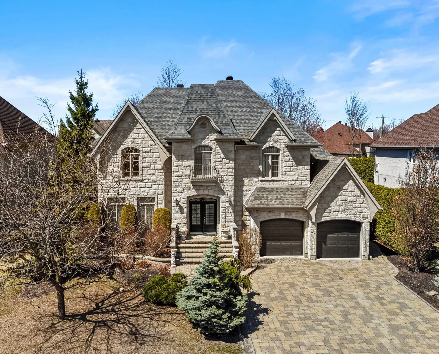 Two-story stone house with a gray roof, two-car garage, and a brick driveway on a sunny day.
