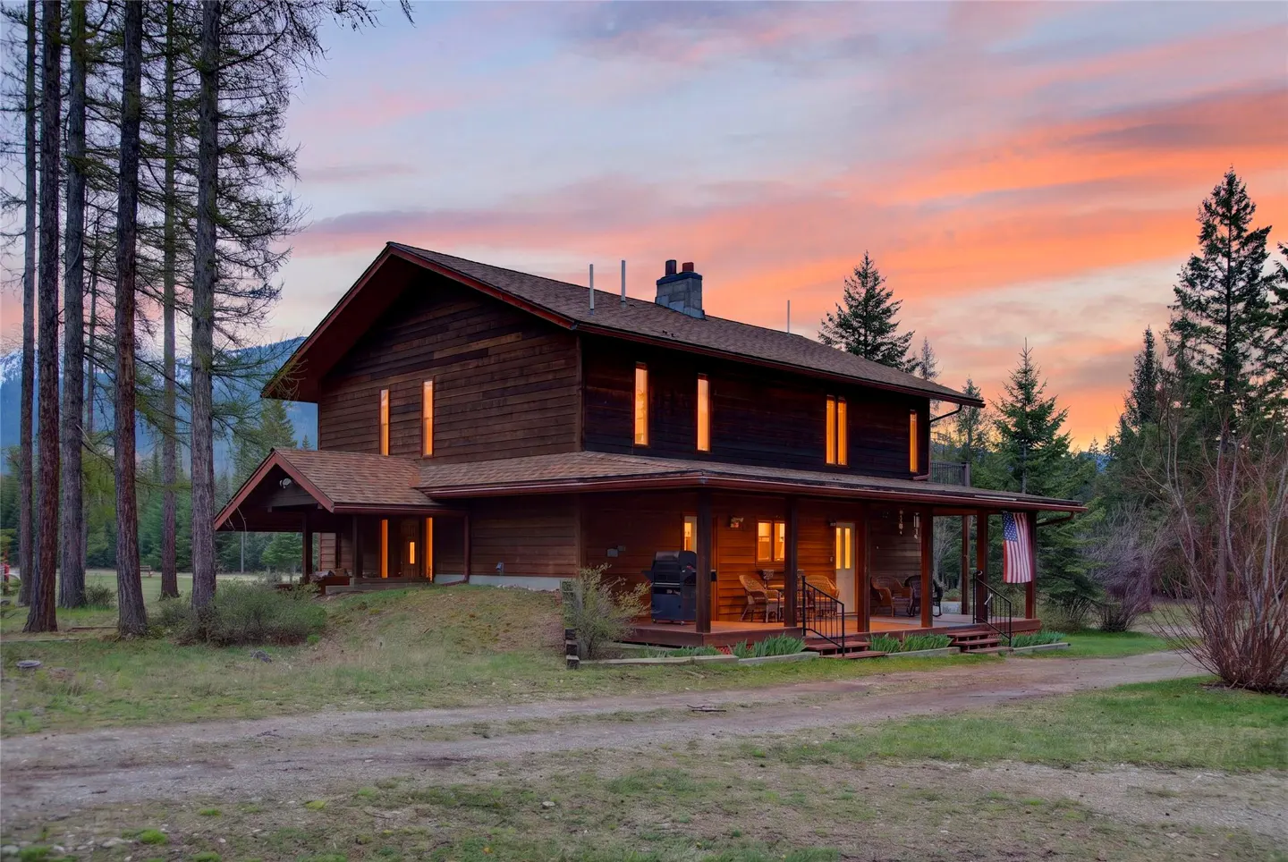 Two-story brown house with a porch and American flag, set against a sunset sky and surrounded by trees.