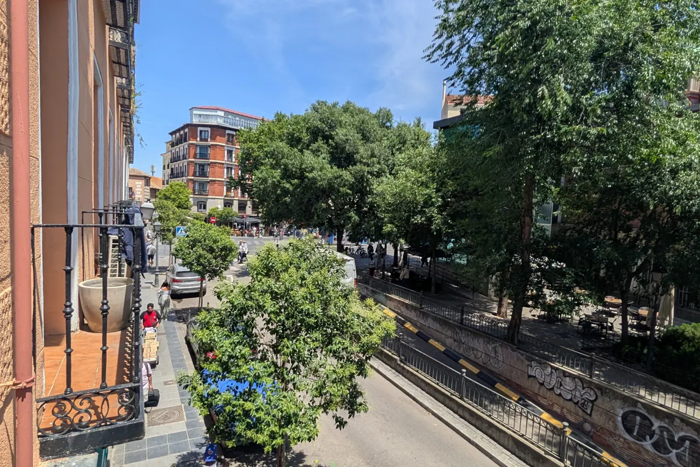 View from a balcony overlooking a street with trees, buildings, and people walking on a sunny day.
