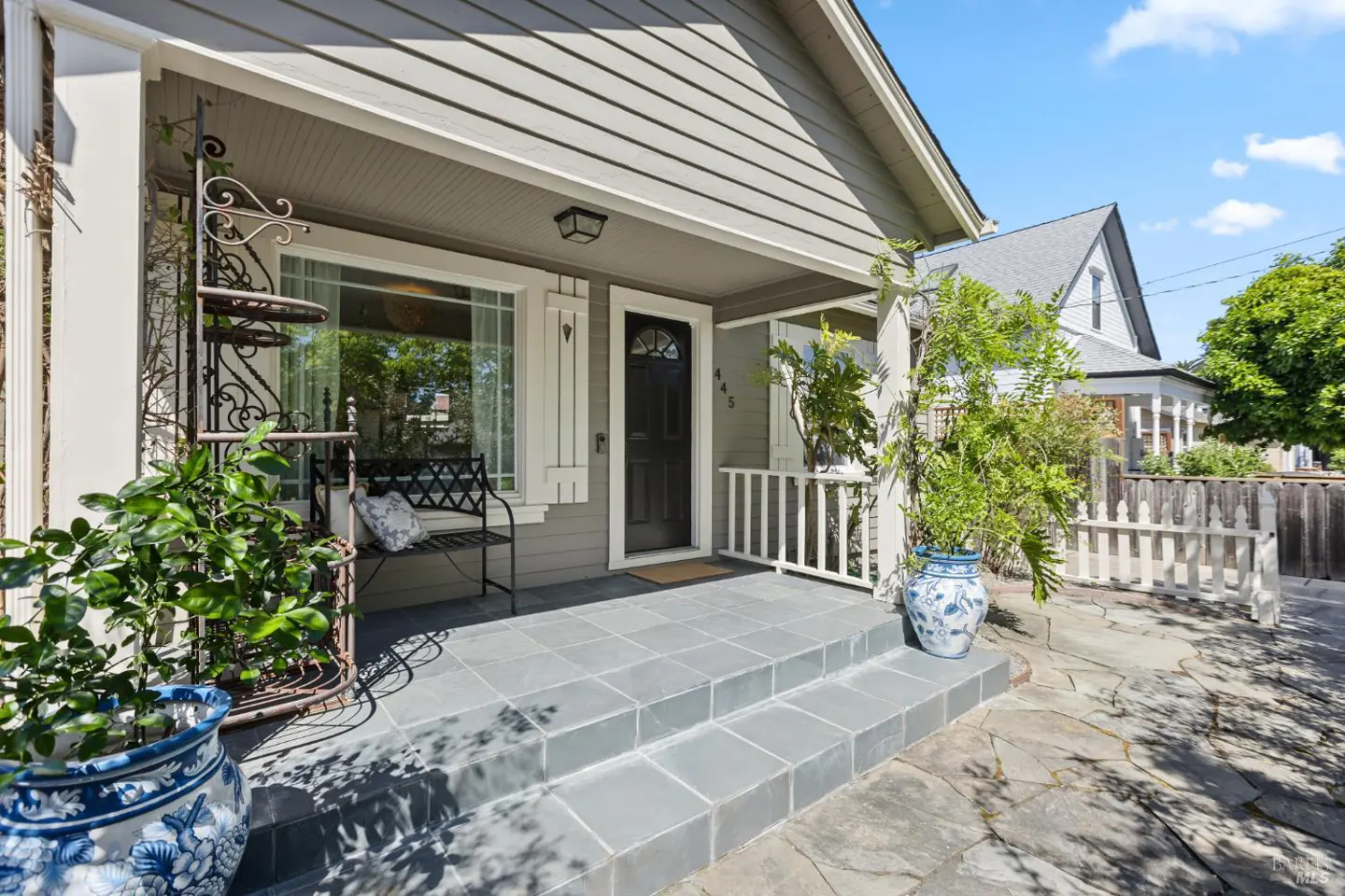 Exterior view of a gray house with a black door, white trim, and a tiled porch with plants and a bench.