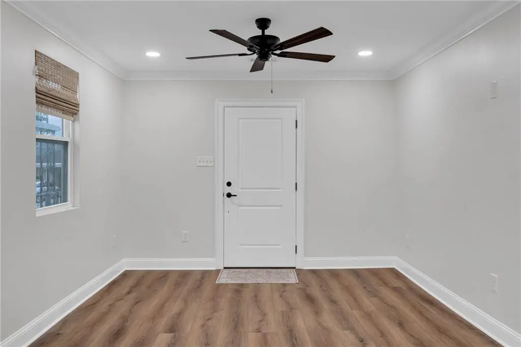 A bright room with wood floors, white walls, and a white door. A dark ceiling fan hangs above, and a window with a woven shade is to the left.