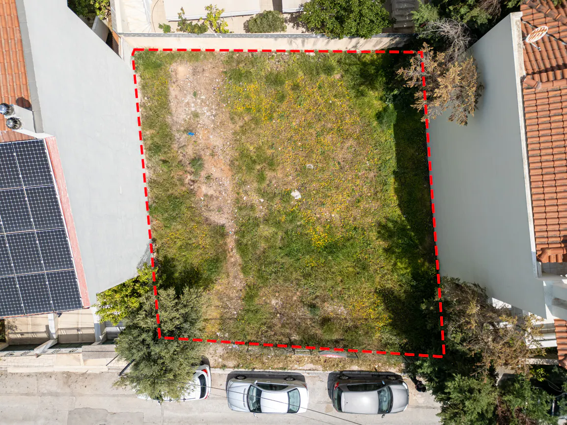 Aerial view of a vacant lot with a red dashed border, surrounded by houses and parked cars.
