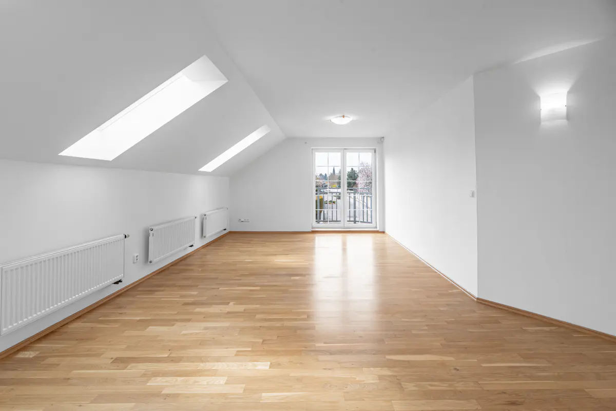 Bright, empty attic room with hardwood floors, white walls, skylights, and a balcony door. Radiators line the wall.
