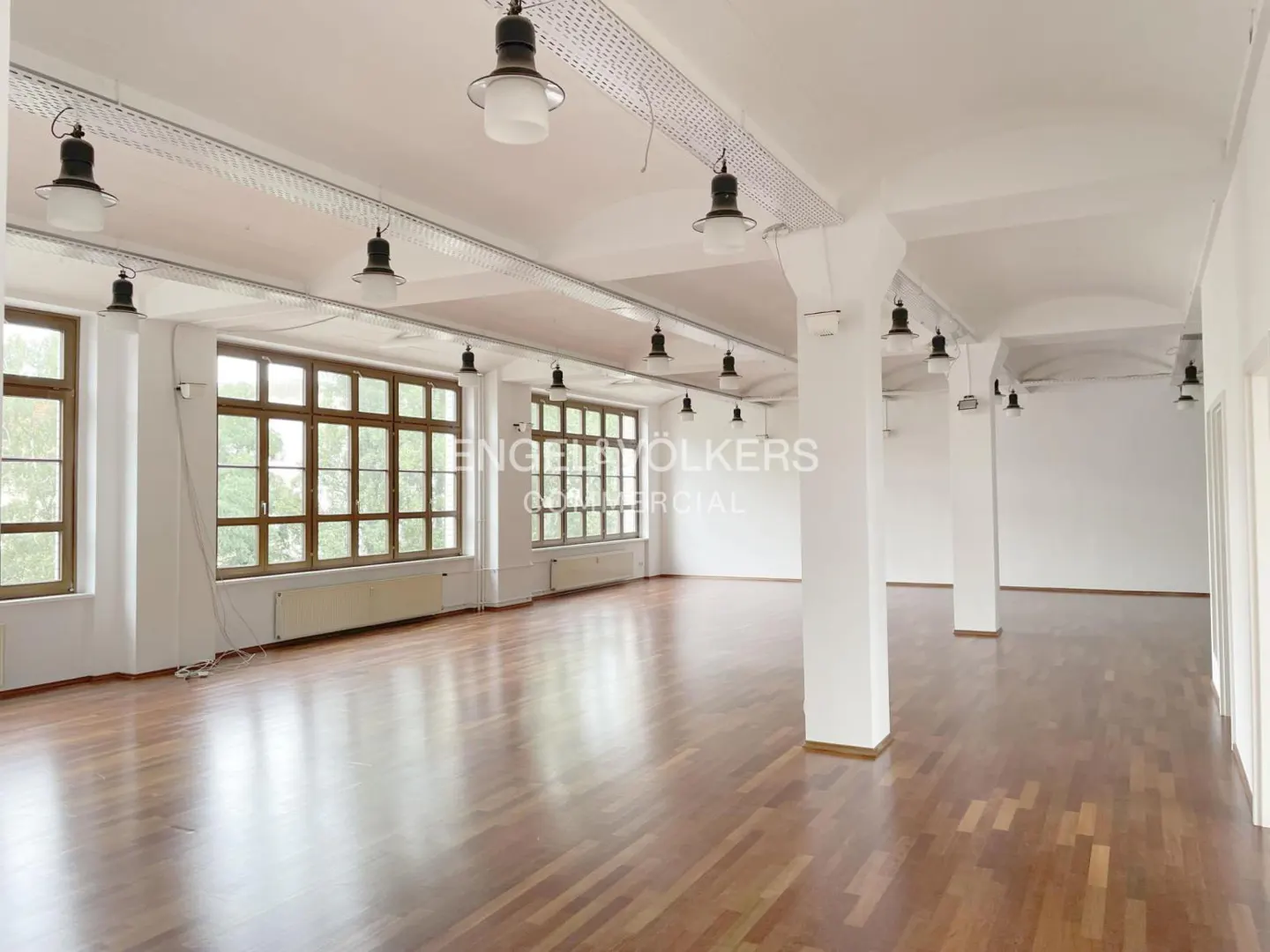Bright, empty commercial space with wood floors, white walls, columns, and large windows. Industrial-style lighting fixtures hang from the ceiling.