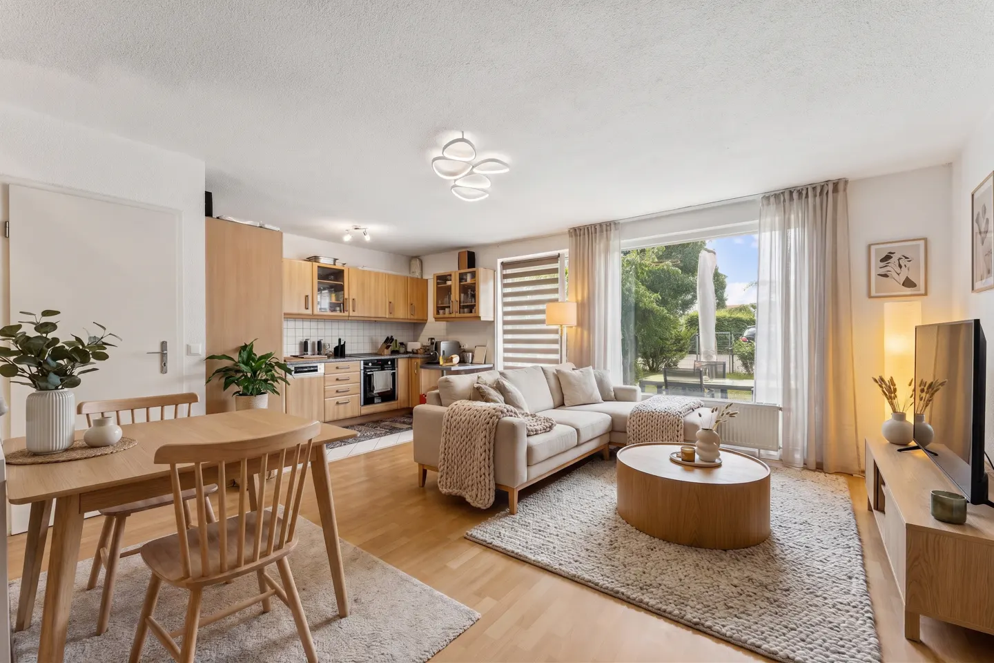 Open-concept living space with light wood floors, cabinets, and furniture. A beige sofa faces a large window with a view of greenery.