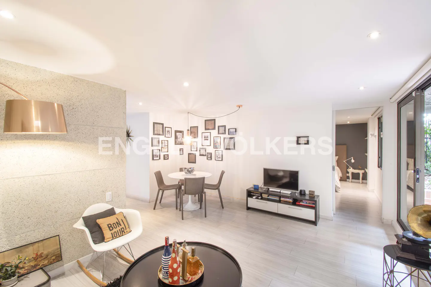 Bright living room with white walls, light wood floors, and modern furniture. A dining table sits near a gallery wall of framed photos.