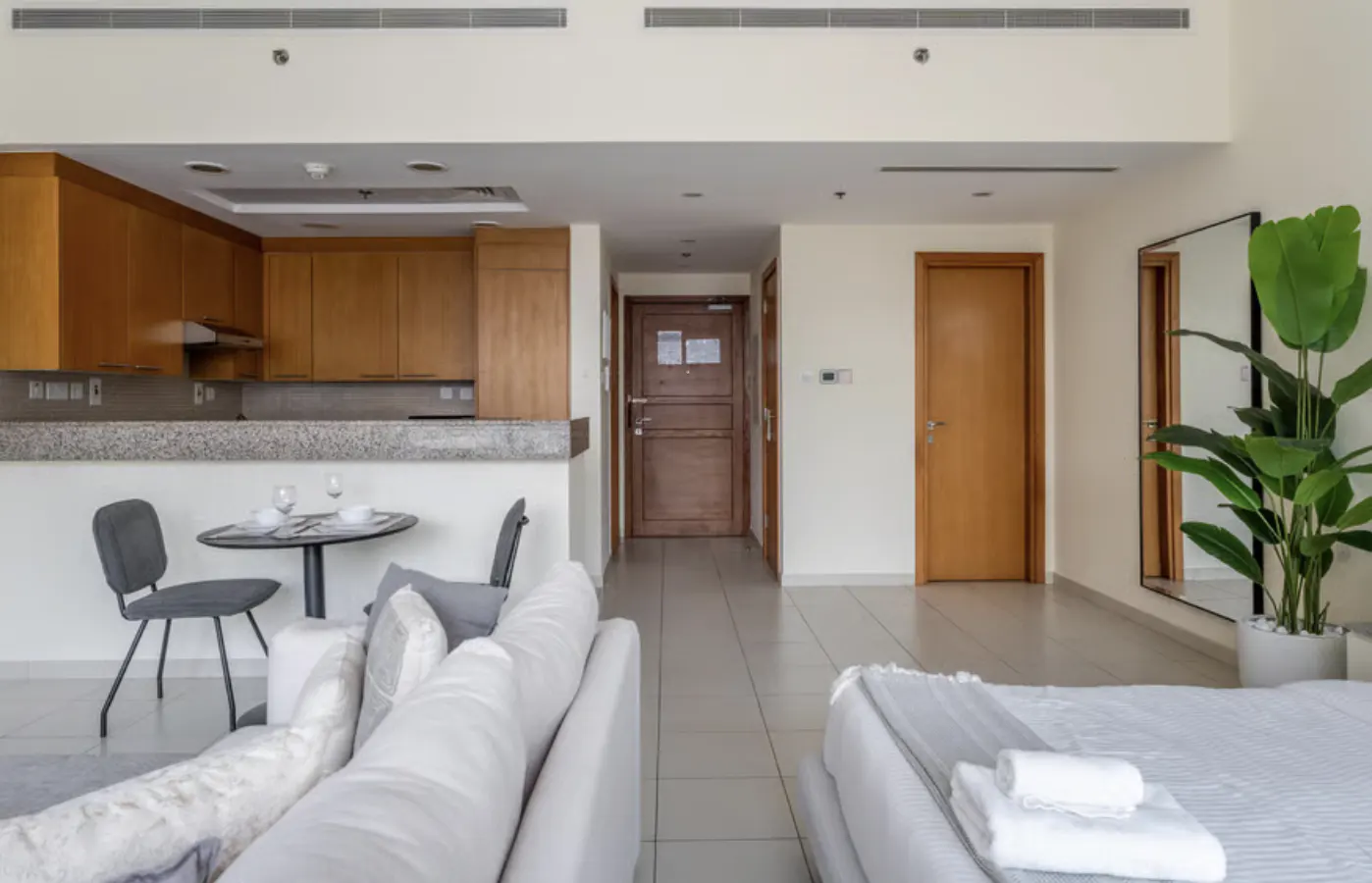 Open-concept apartment with a kitchen, dining table, sofa, and bed. Neutral tones with wood accents and a potted plant.
