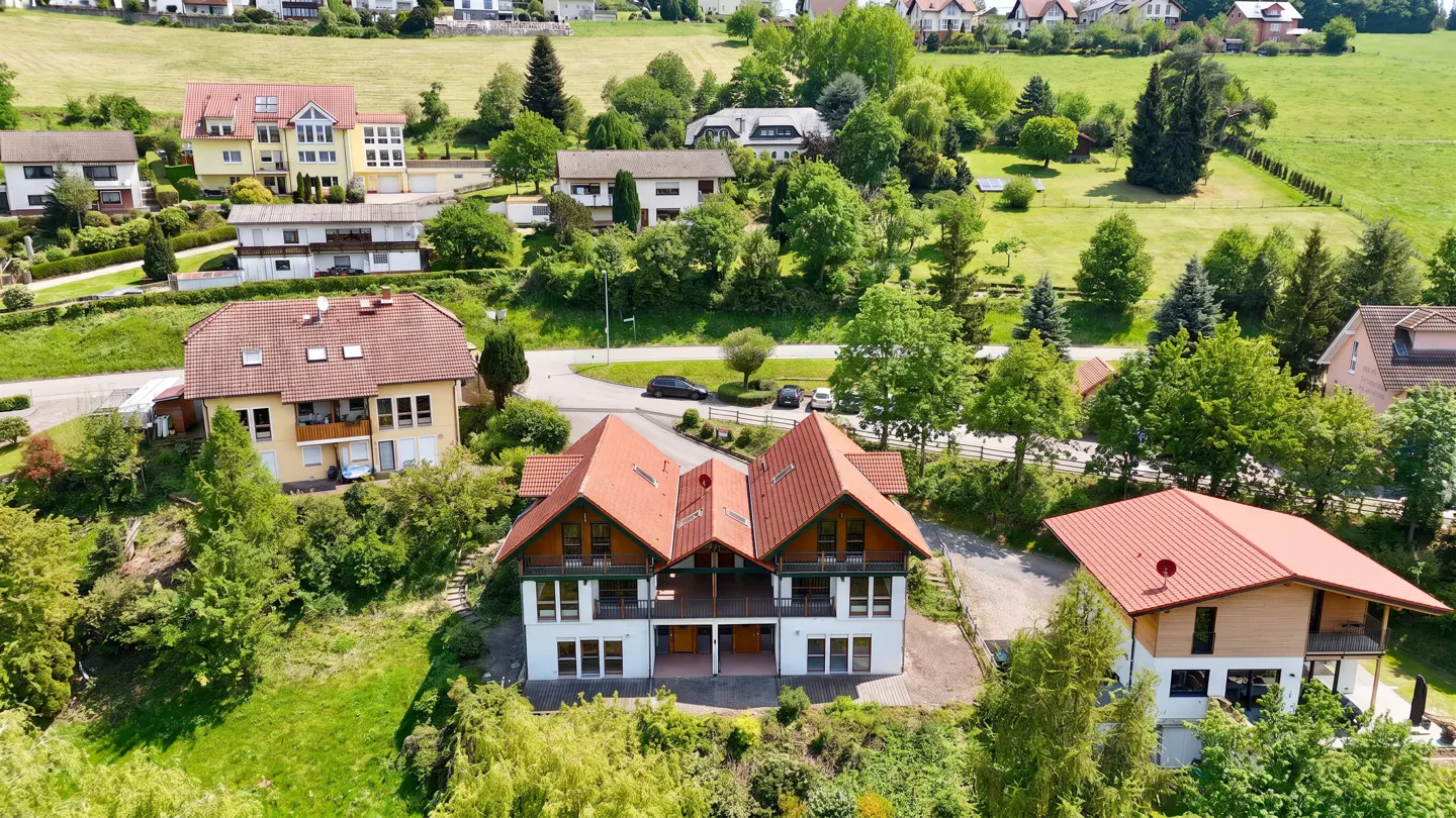 A high angle shot of a white two-story house with a red roof, surrounded by green trees and other houses in the background.