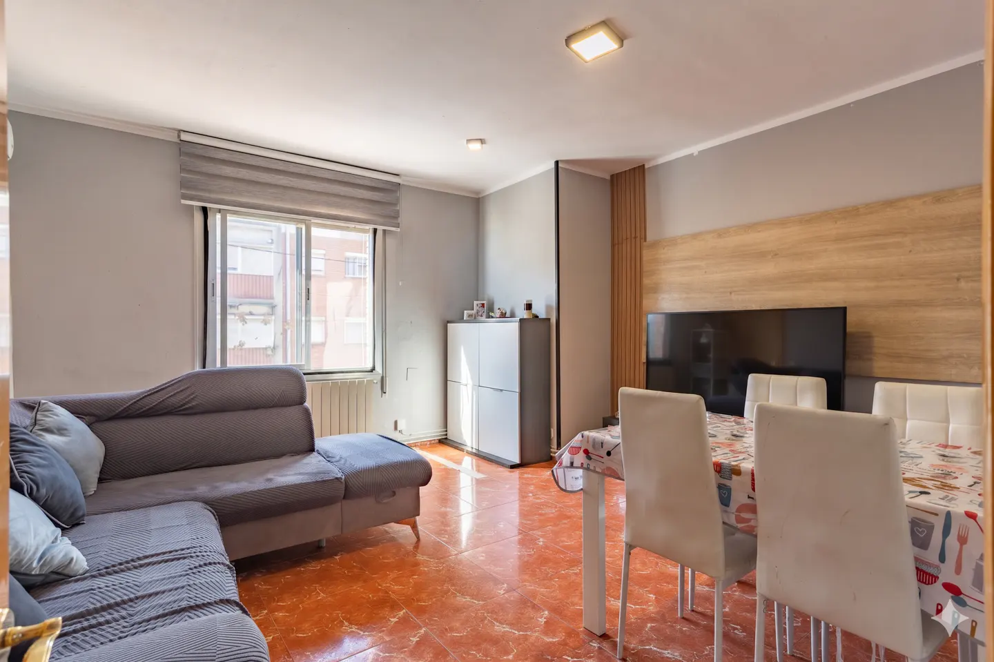 Living room with grey sectional sofa, dining table with white chairs, and a TV mounted on a wood-paneled wall.