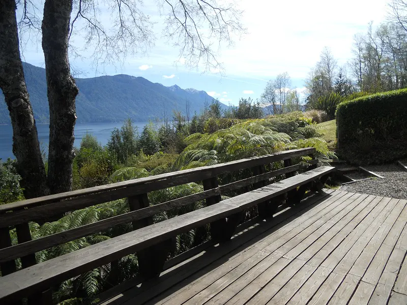 Wooden deck with bench overlooking a lake and mountains. Ferns and trees fill the landscape.