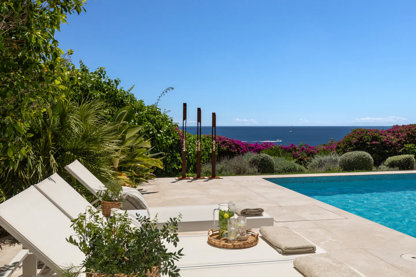 Luxury poolside view with lounge chairs, a drink tray, and ocean view. Lush greenery and blue sky create a serene backdrop.