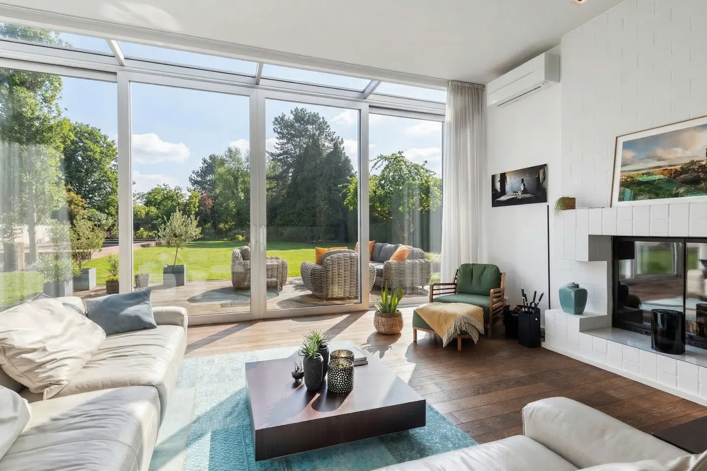 Bright living room with white sofas, a dark wood coffee table on a blue rug, and a fireplace. Large windows overlook a green lawn and patio furniture.
