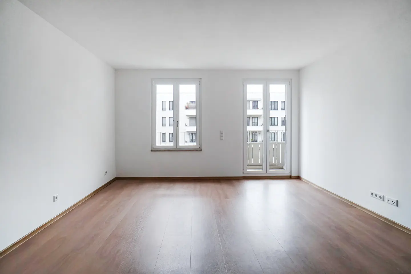 Empty room with white walls, brown wood floor, and two white windows. Outside, a building is visible.