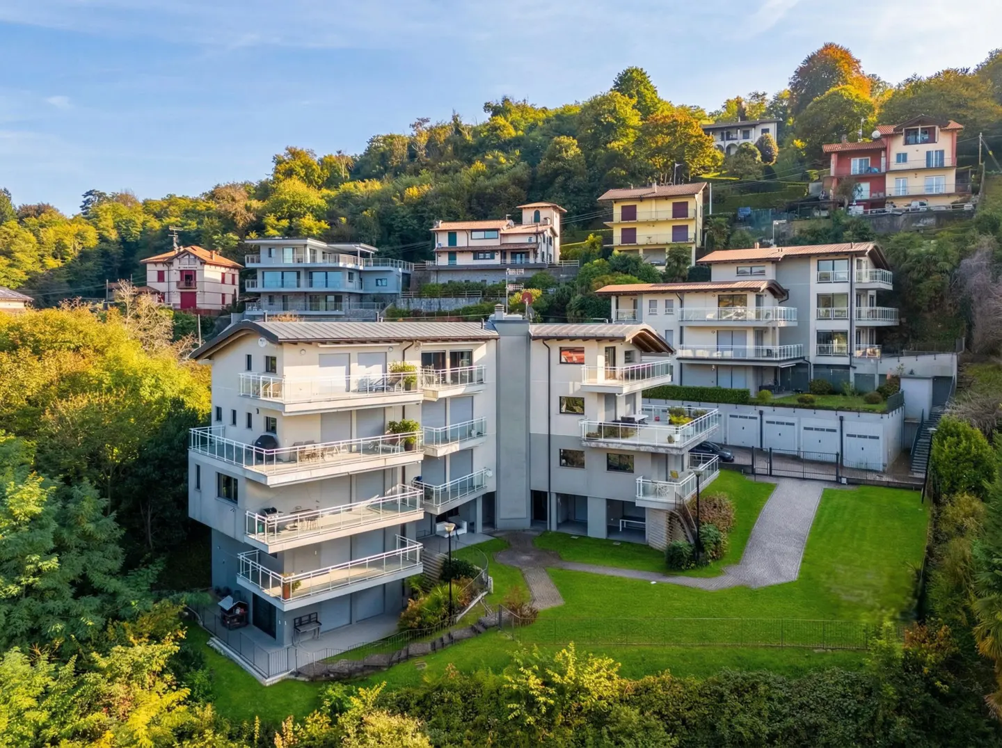 View of modern, multi-story apartments with balconies on a green hillside under a blue sky.