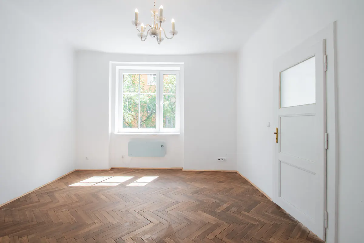Bright, empty room with white walls, herringbone wood floor, window with trees outside, chandelier, and white door.