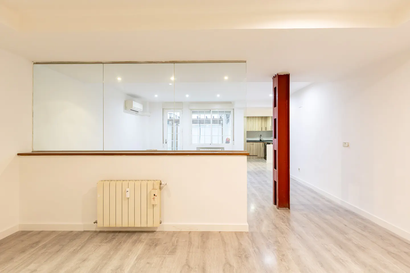 Bright, empty room with wood floors, white walls, and a red support beam. A half wall with a glass panel separates the space from a kitchen area. A radiator is on the wall.