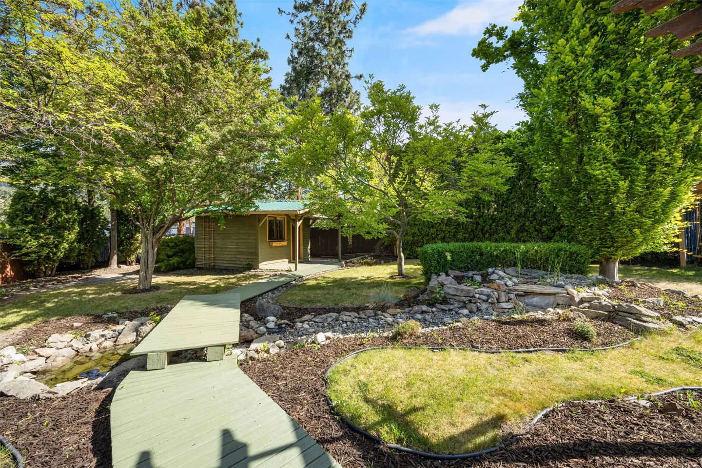 Landscaped backyard with a green wooden walkway over a small stream, leading to a shed and patio area. Trees and shrubs surround the yard.