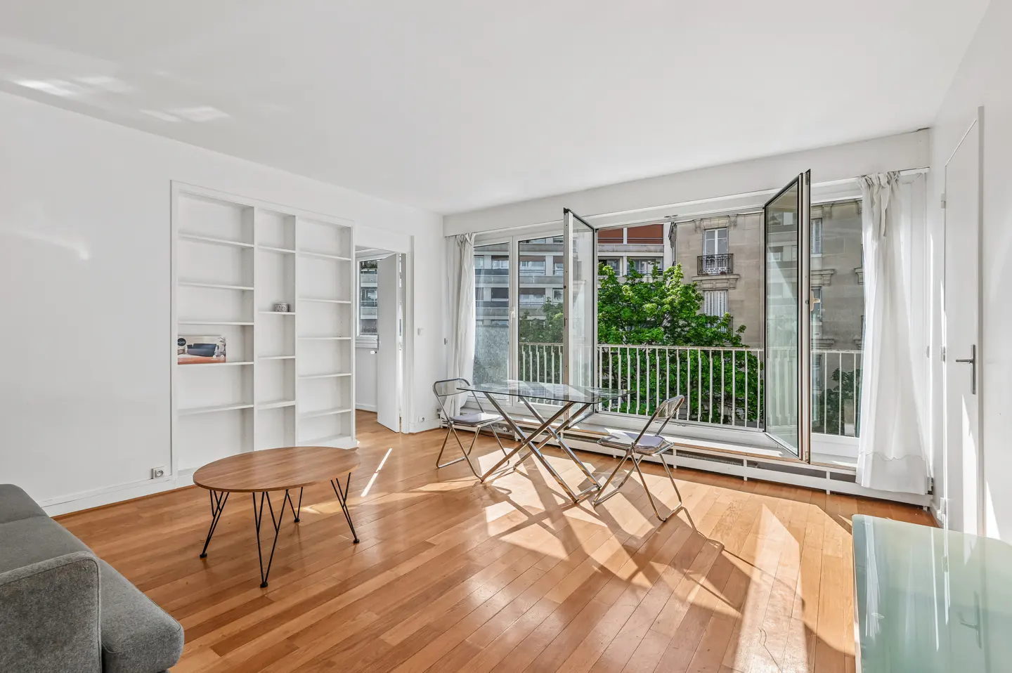 Bright living room with wood floors, white walls, and built-in shelves. Balcony with table and chairs overlooks a city view.
