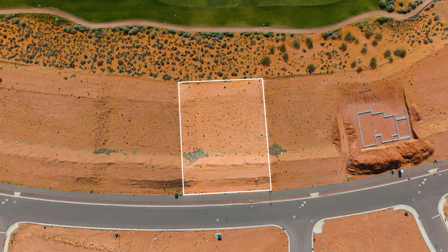 Aerial view of a vacant, rectangular lot outlined in white, next to a road and a house foundation. The land is reddish-brown.