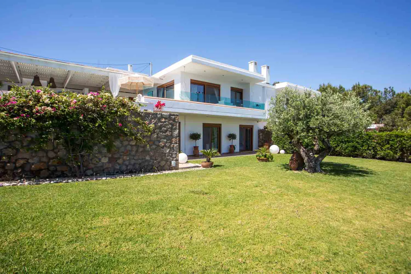 Two-story white house with a green lawn, stone wall, and olive tree under a blue sky.