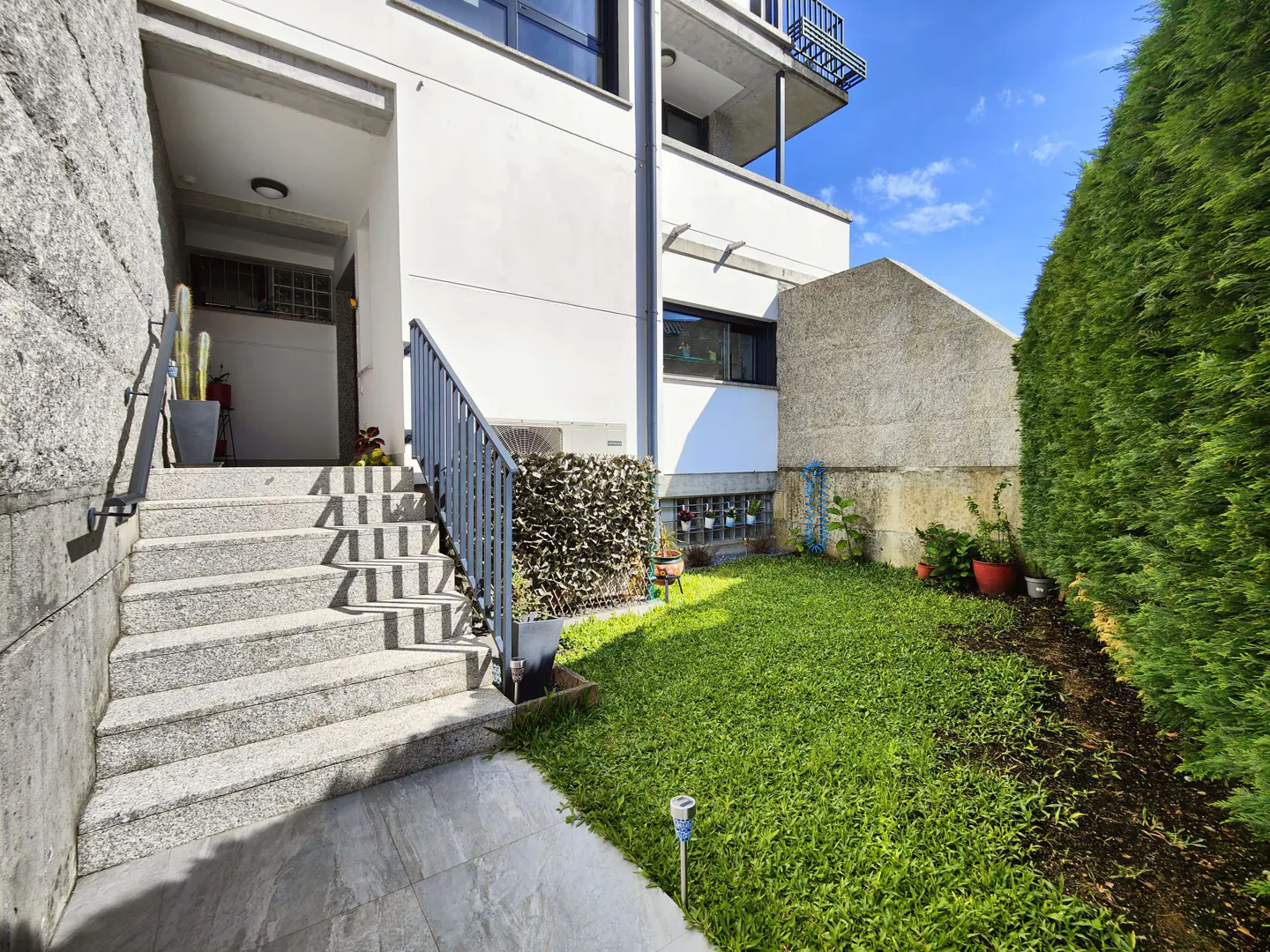 Exterior view of a white building with stone steps leading to the entrance, a small green lawn, and a tall hedge.