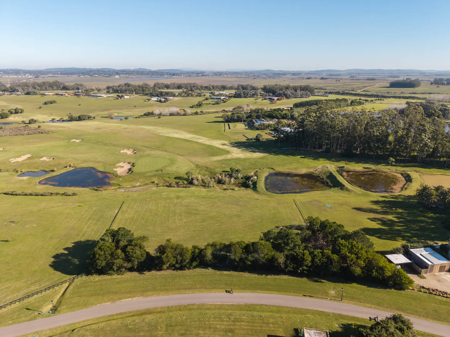 Aerial view of a vast green landscape with ponds, trees, and a paved road in the foreground under a clear blue sky.