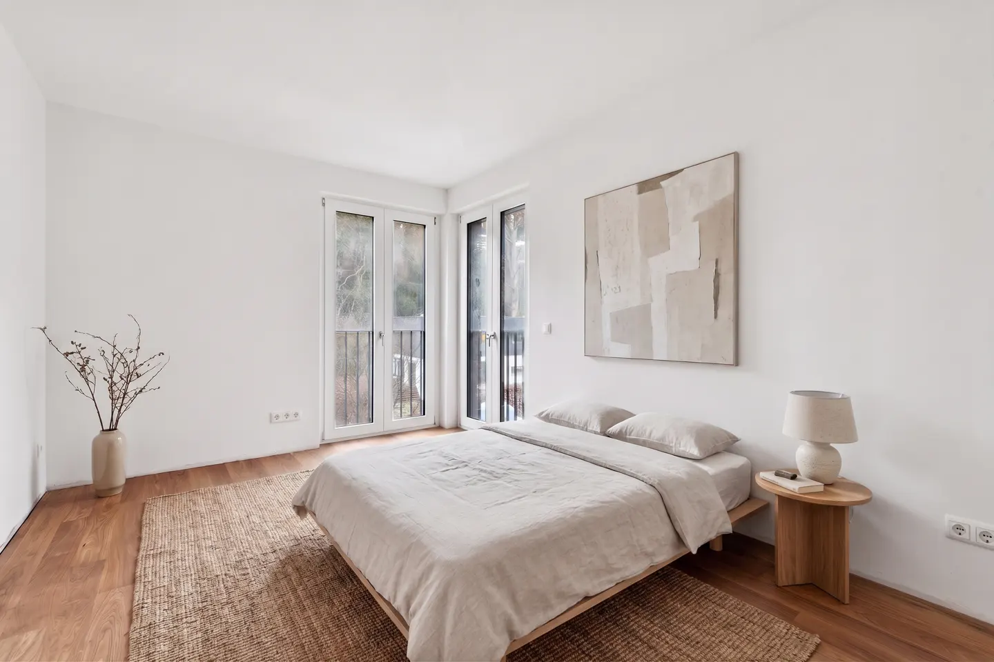 Bright bedroom with white walls, wood floors, and a jute rug. A bed with neutral linens sits beneath an abstract painting. French doors lead to a balcony.