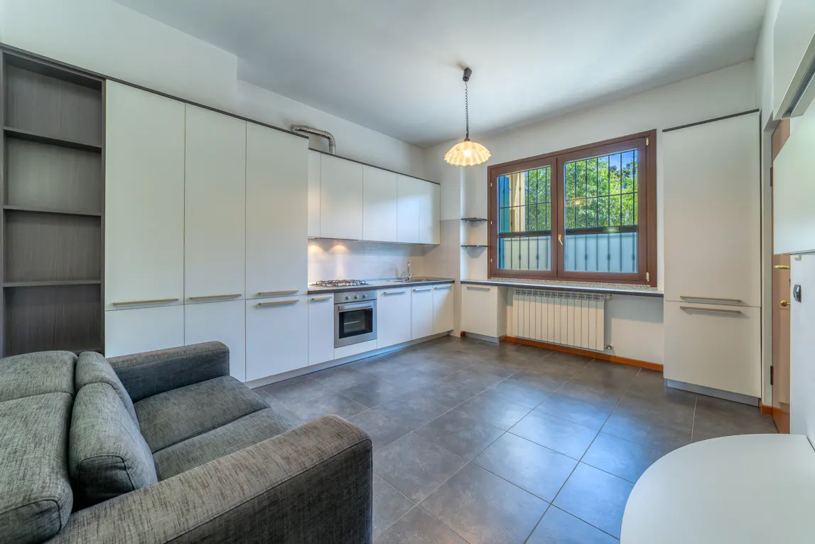 A modern kitchen with white cabinets, gray tile floor, and a gray sofa. A window with a brown frame overlooks a green outdoor space.