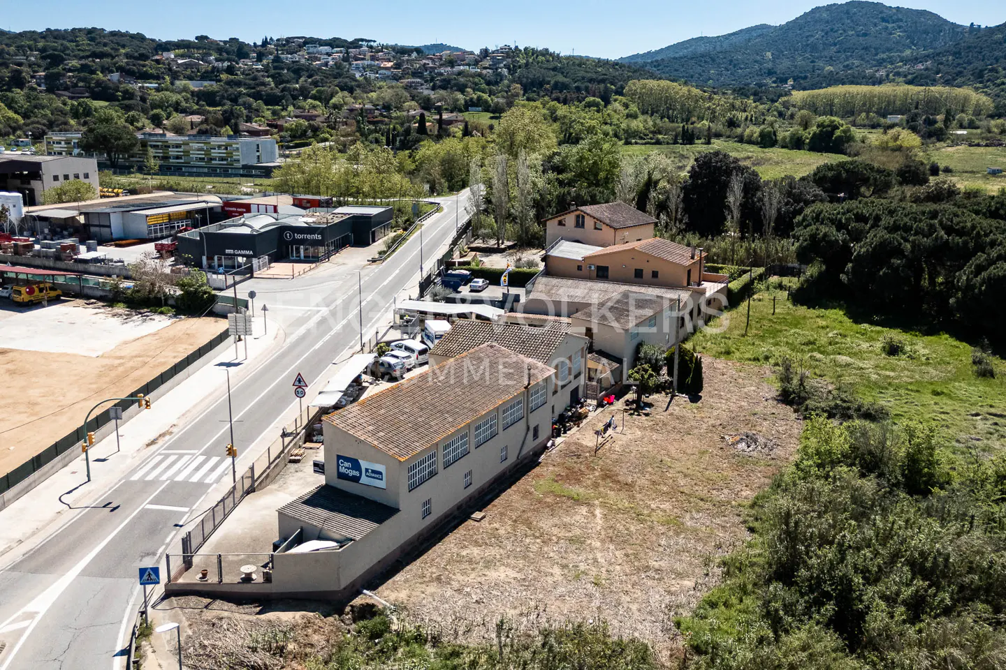 Aerial view of a commercial building with a brown tile roof, next to a road with parked cars and green trees in the background.