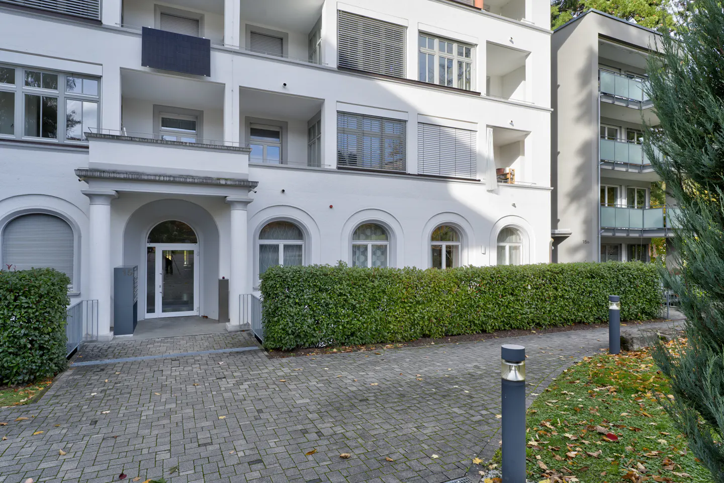 Exterior of a white apartment building with arched windows, a green hedge, and a brick walkway.