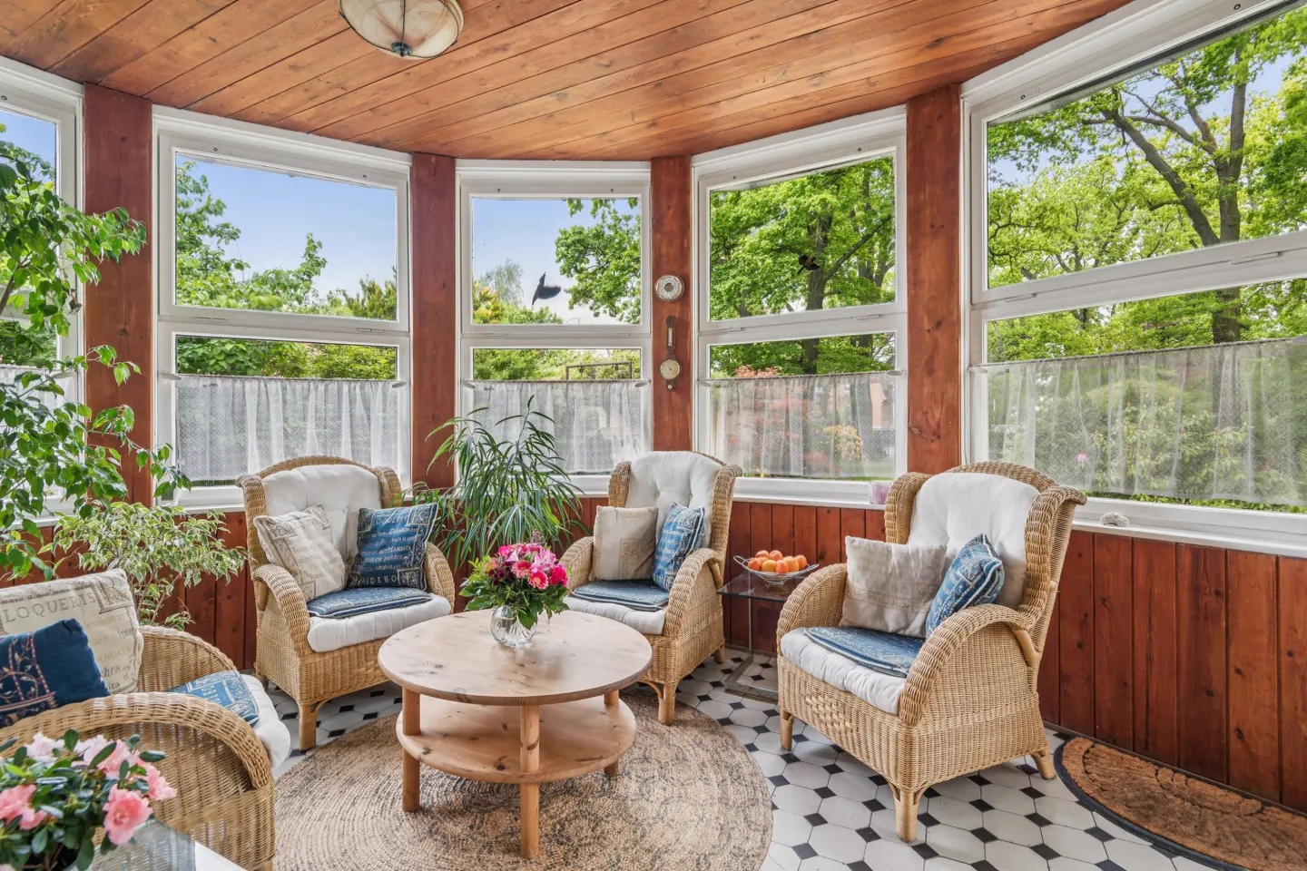 Sunroom with wicker chairs, wood walls, and large windows overlooking green trees. A round table holds flowers.