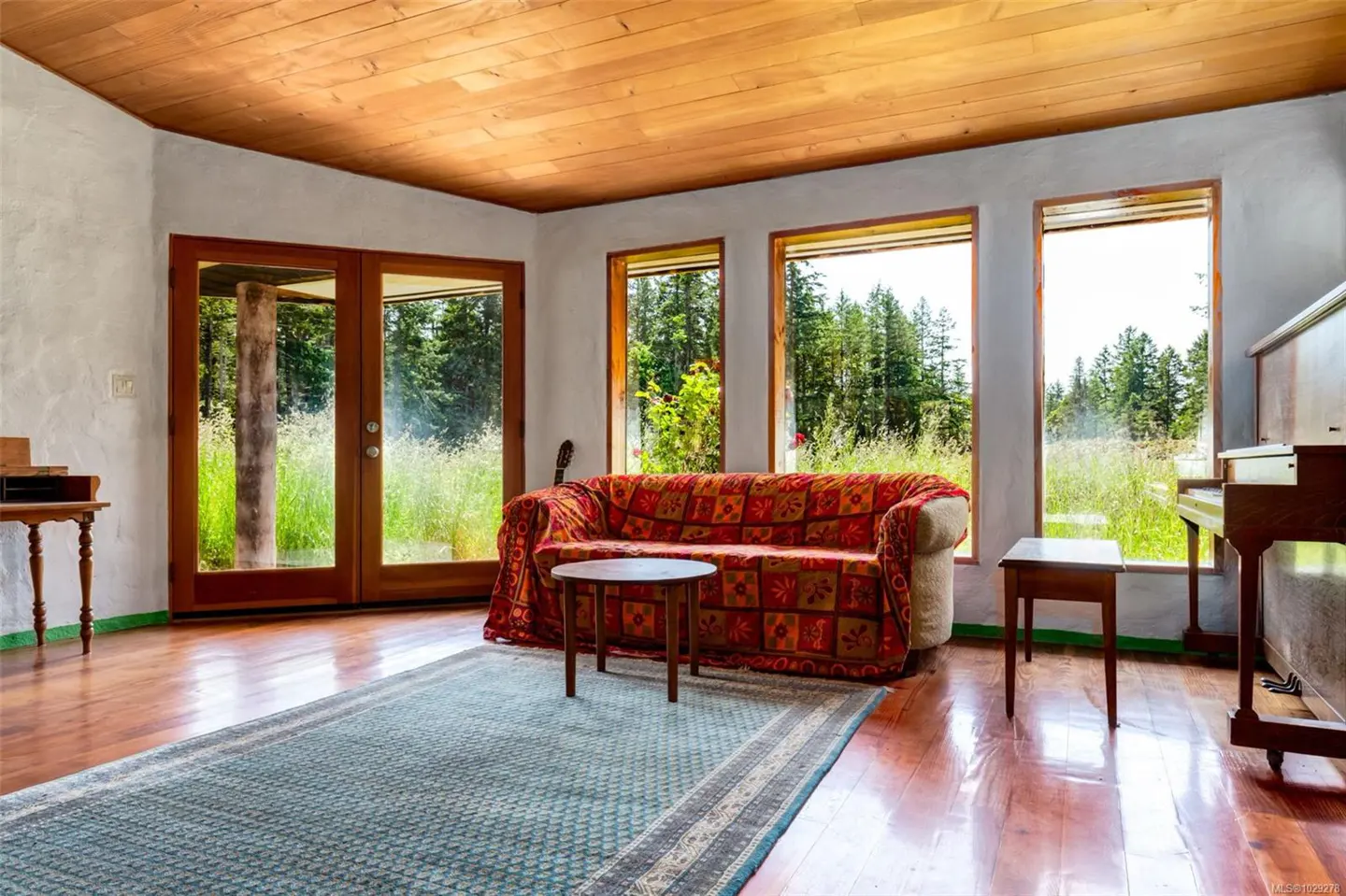 Living room with wood floors, a patterned sofa, and a blue rug. Large windows offer views of trees and greenery. A piano sits to the right.