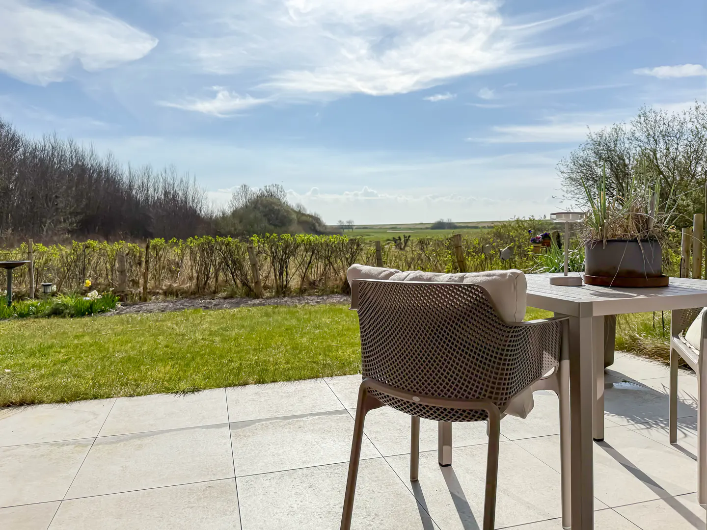 Outdoor patio with a table, chair, and potted plant overlooking a green lawn and distant field under a blue sky with white clouds.
