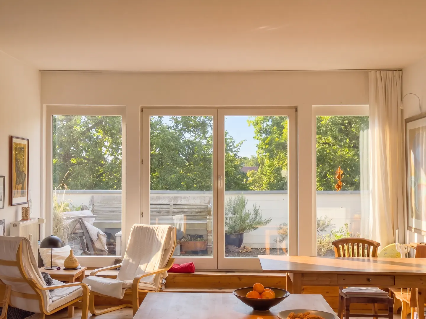 Bright living room with two armchairs, a wooden table with fruit, and a large window overlooking a balcony with green trees.