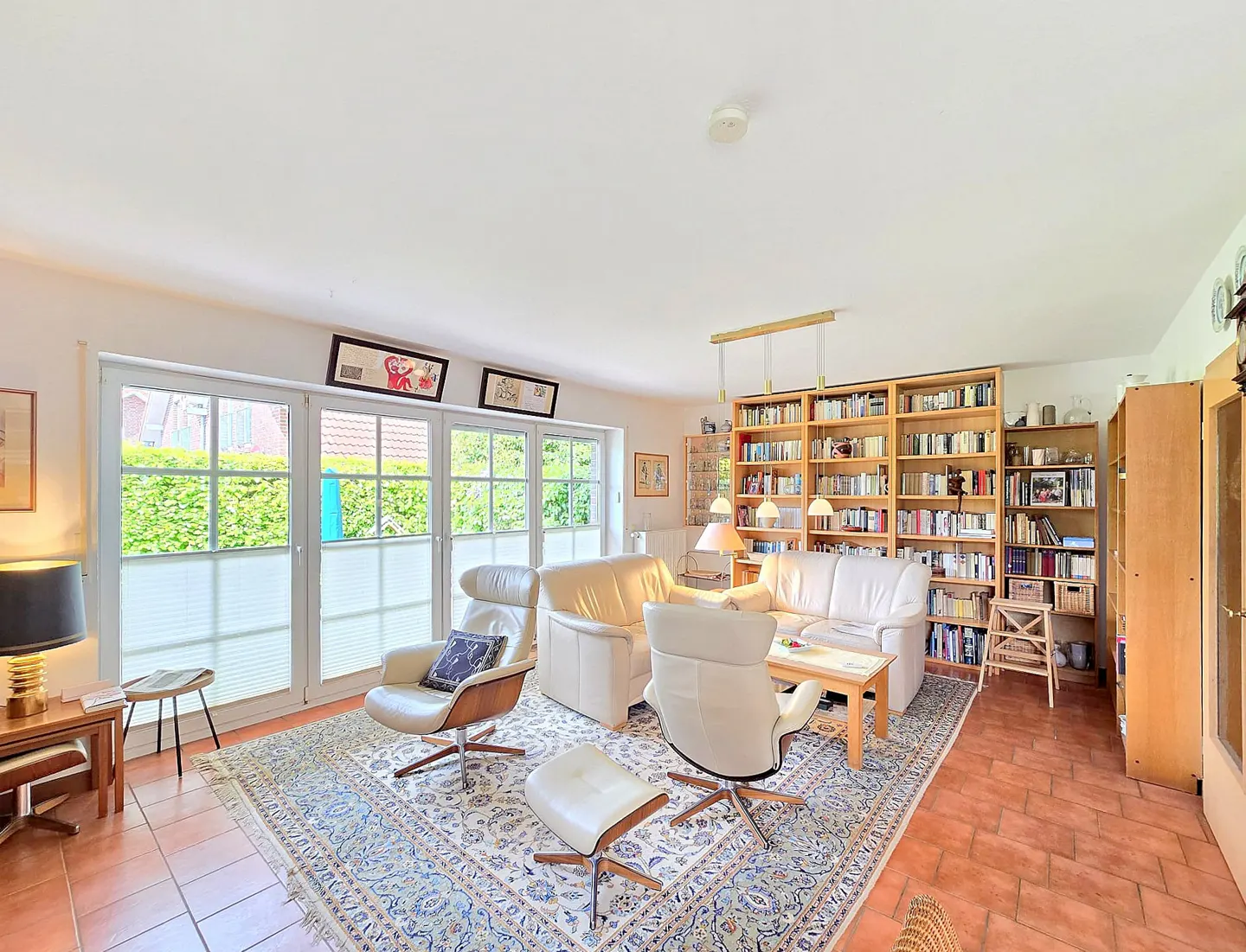 Bright living room with white leather chairs, a sofa, and floor-to-ceiling bookshelves filled with books. A blue patterned rug covers the terracotta tile floor.
