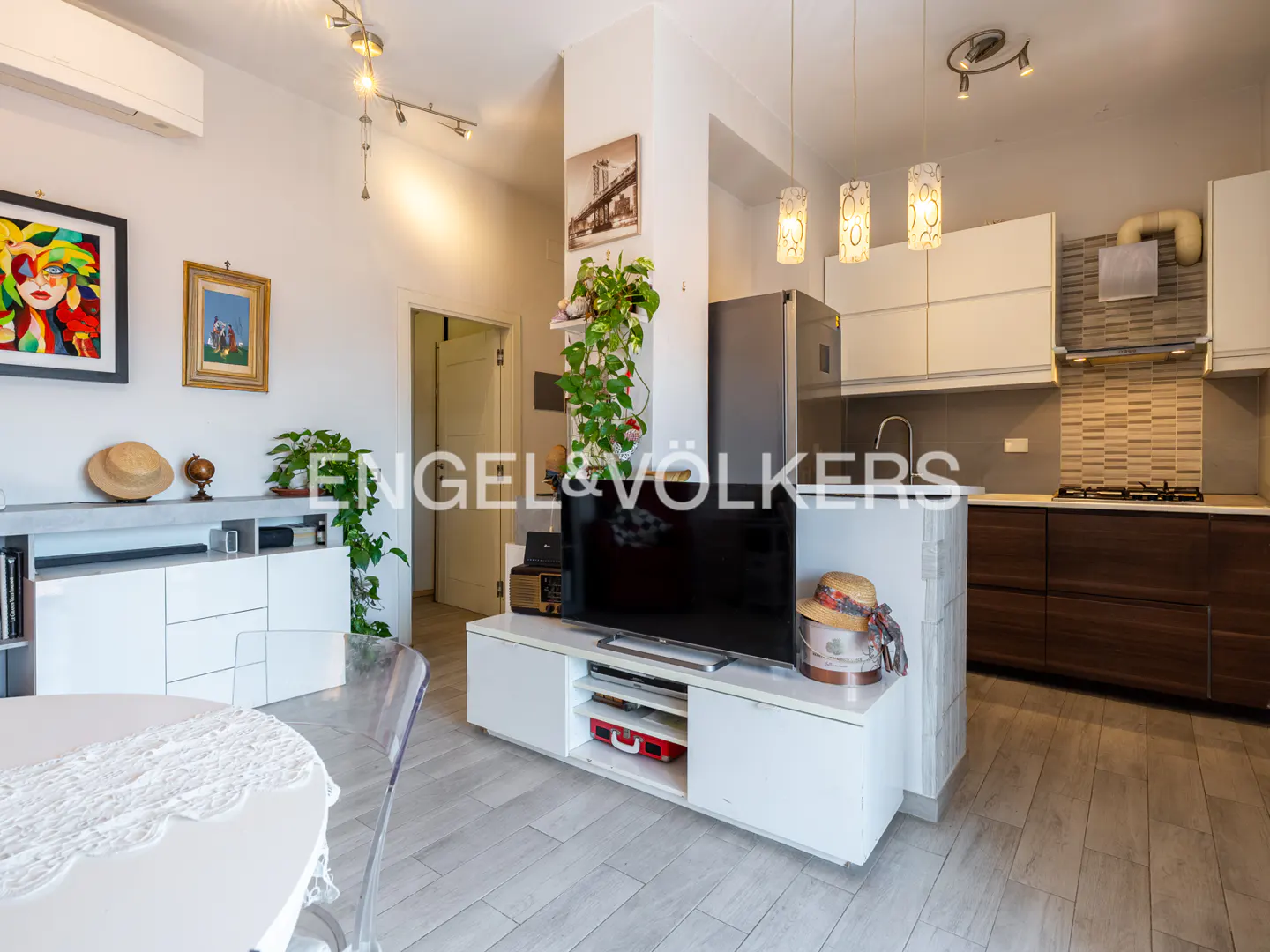 A bright, modern apartment interior with white walls, light wood floors, and an open kitchen. A TV sits on a white console. Artwork and plants add color.