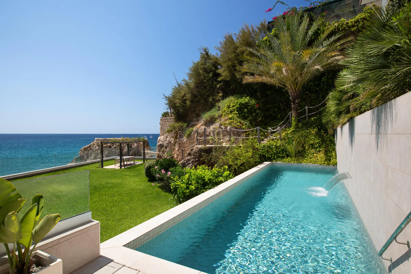 Outdoor pool with waterfall feature, green lawn, and ocean view under a clear blue sky. Lush greenery surrounds the pool area.