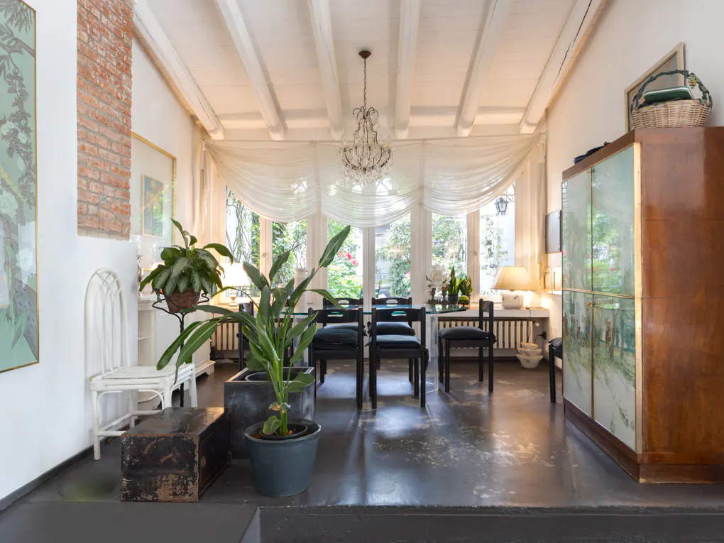 Bright dining room with a glass table, black chairs, and a chandelier. Sheer white curtains adorn the windows. Plants and a wooden cabinet add to the decor.