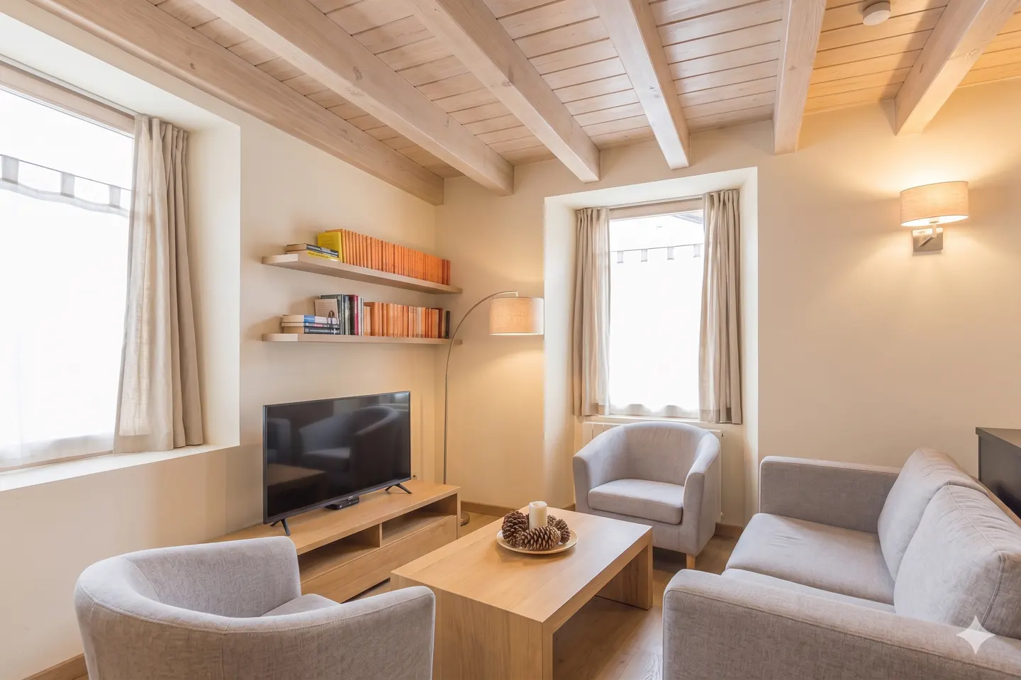 Living room with gray sofa and chairs, wood ceiling beams, TV, and bookshelves. A coffee table with pine cones sits in the center.