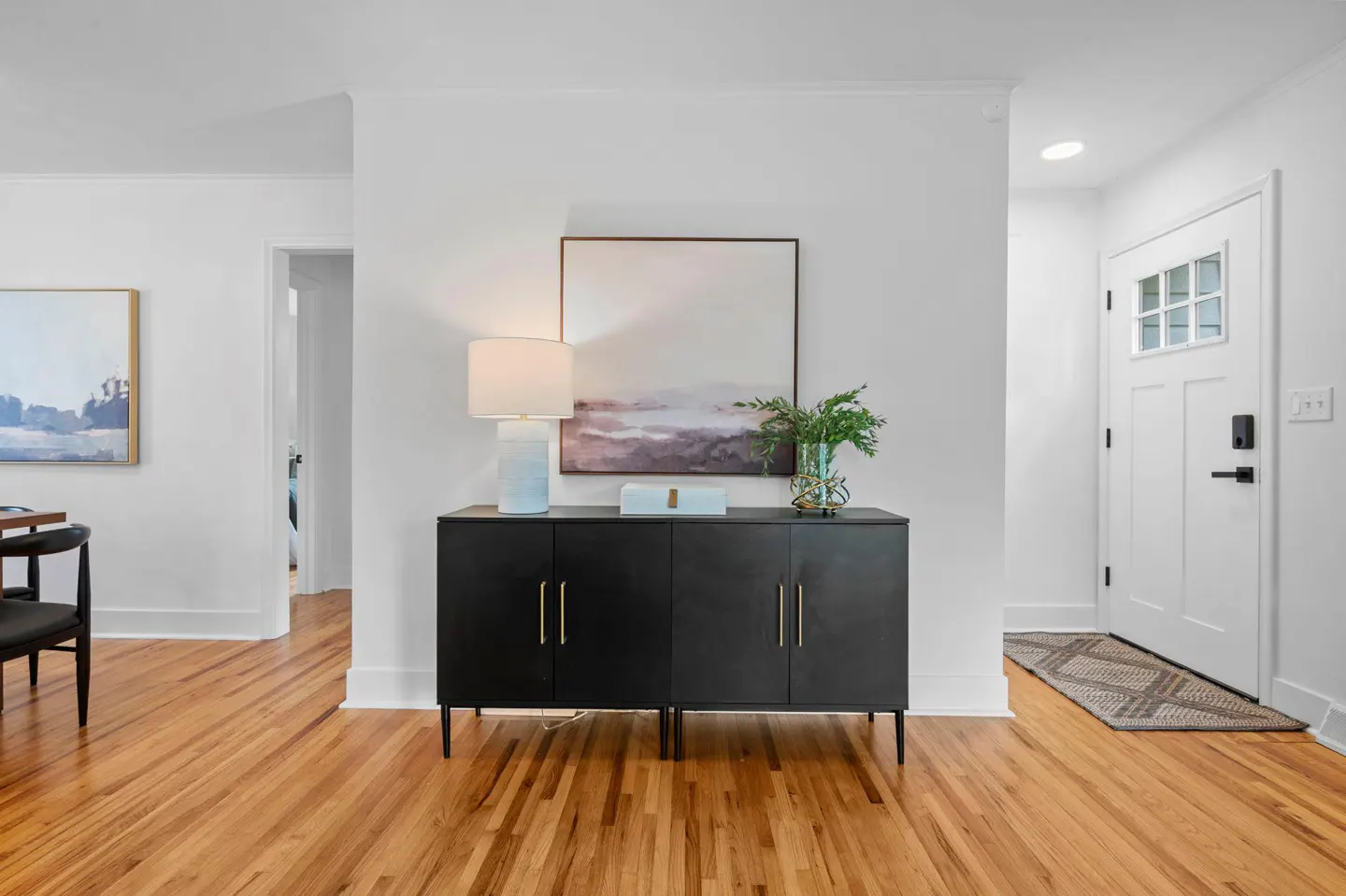 Black sideboard with lamp, decor, and abstract art on a white wall in a bright entryway with hardwood floors.