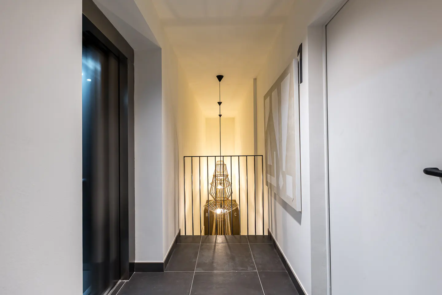 Hallway with dark tile floor, white walls, and a black metal railing overlooking a lower level with a modern light fixture.