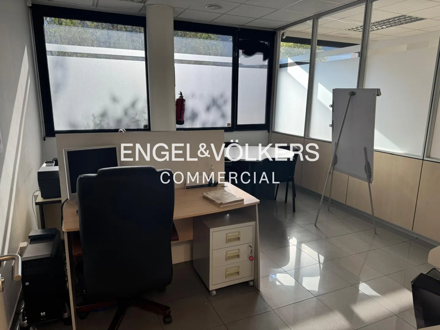 Office space with desk, computer, chair, and whiteboard. Windows with frosted glass provide natural light. Engel & Völkers Commercial logo visible.