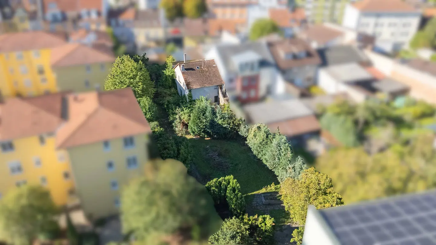 Aerial view of a small white house with a red tile roof, surrounded by lush green trees and grass. Other buildings are blurred in the background.