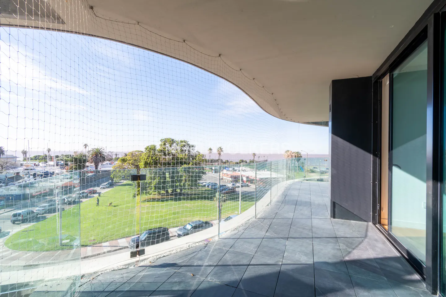 Balcony view with glass railing and safety net overlooking a park, street, and ocean on a sunny day.