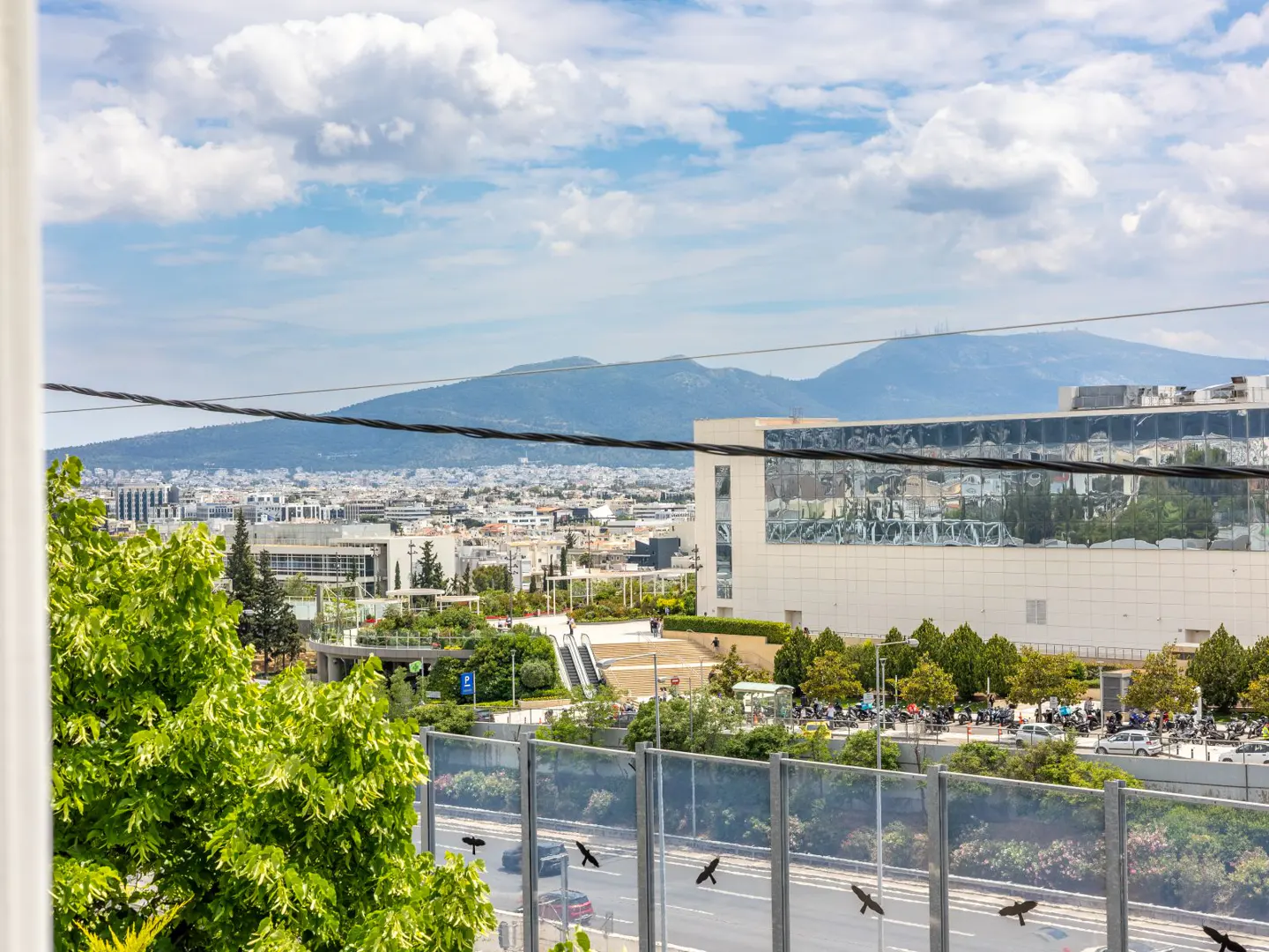 View of Athens, Greece, with mountains in the distance under a cloudy sky. A modern building with reflective windows is visible. Green trees and a highway are in the foreground.